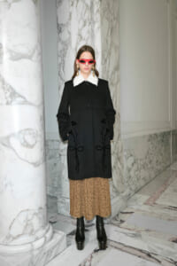 Model wearing a black coat with a white fur collar, leopard-print midi skirt, and black platform boots in a marble hallway.