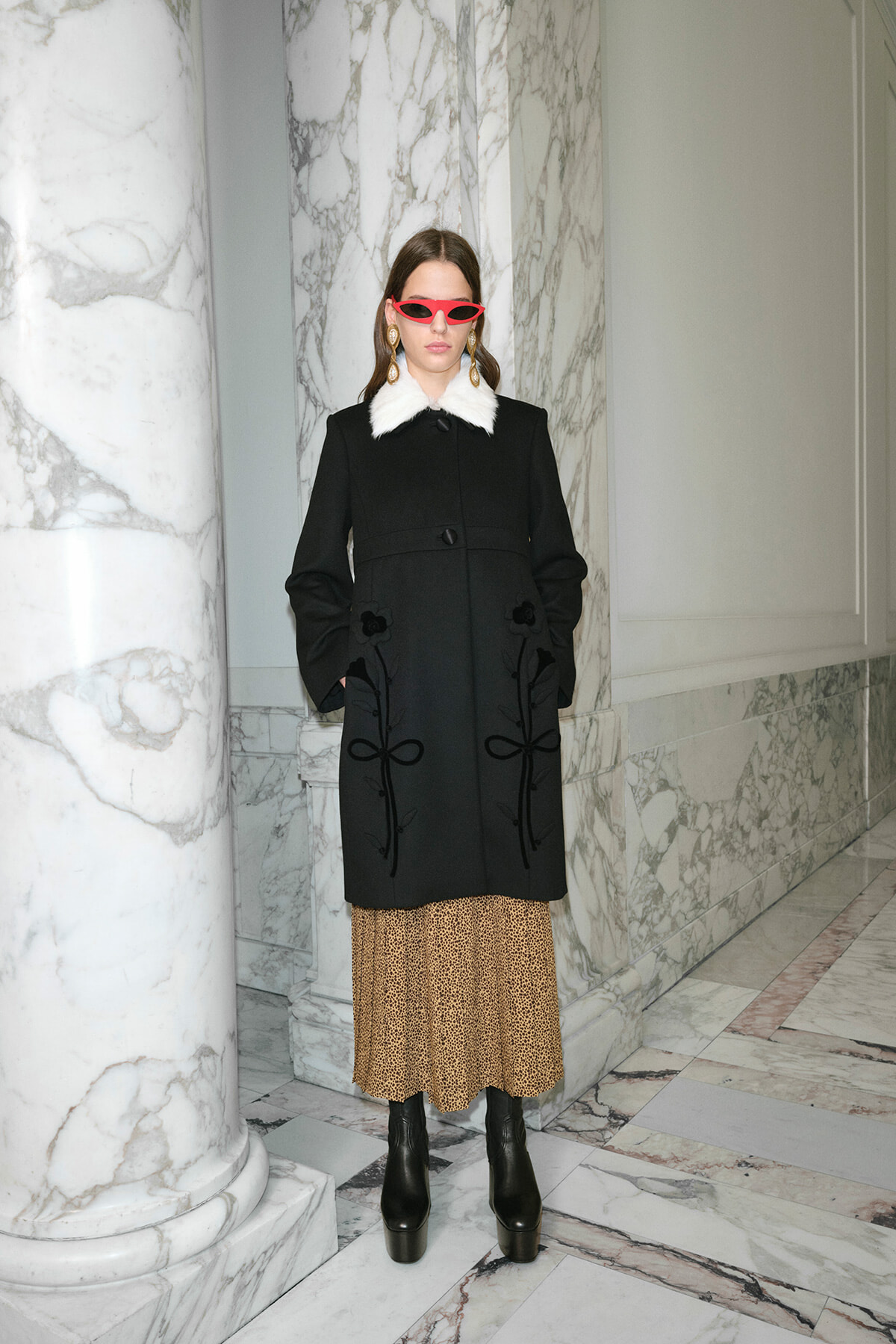 Model wearing a black coat with a white fur collar, leopard-print midi skirt, and black platform boots in a marble hallway.