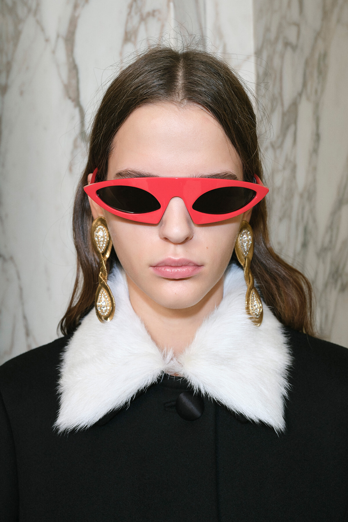 Woman wearing bright red futuristic sunglasses, large gold earrings, and a white fur-collar black coat (fashion portrait).