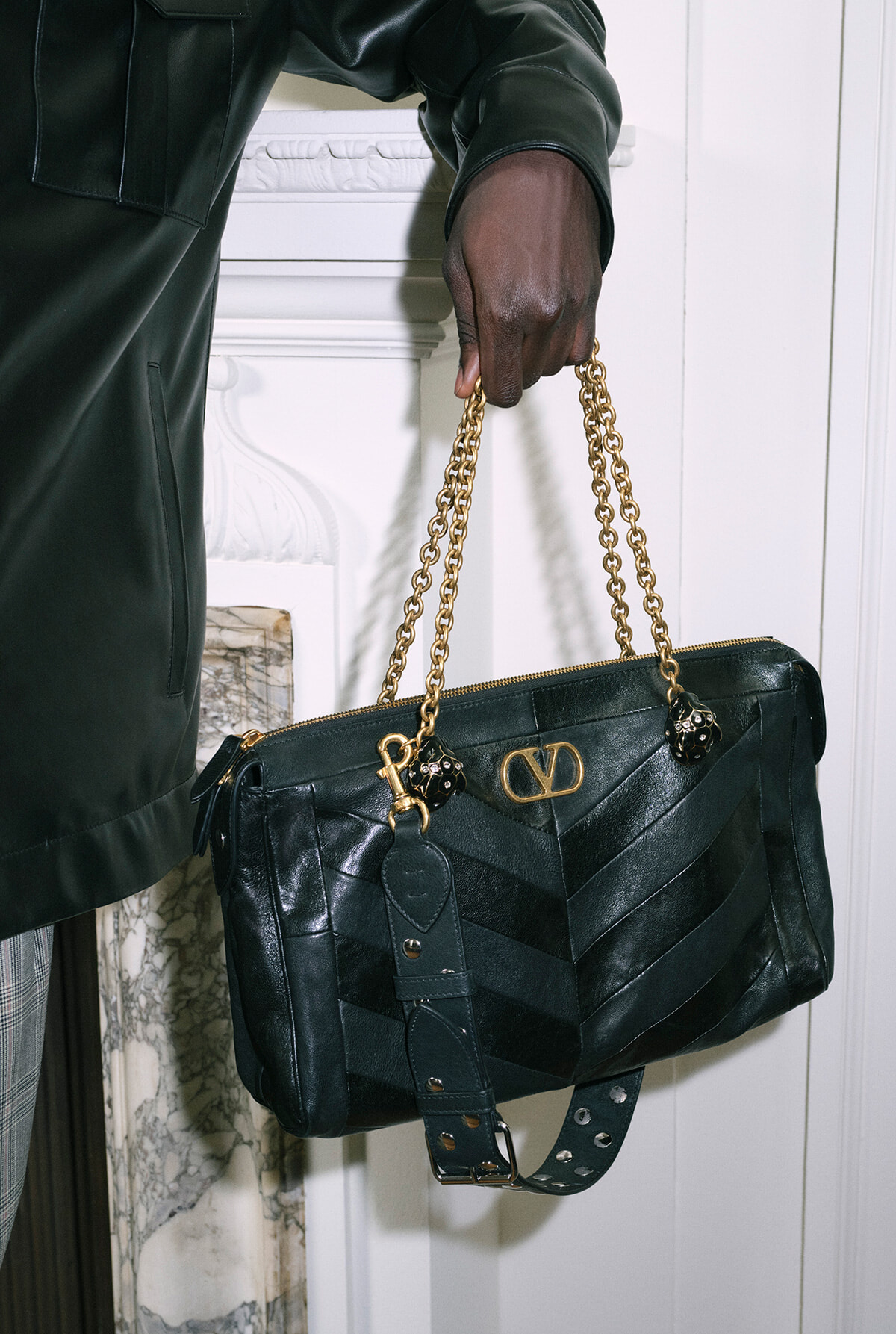 Black quilted leather handbag with gold chain handles being held by a person in a dark jacket on a white background prompt edge.