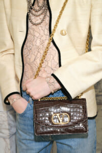 Person wearing a cream blazer with black trim, a pink lace top, layered necklaces, and a brown crocodile-embossed handbag with a gold chain strap.
