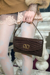 Close-up of a hand holding a dark brown crocodile-embossed handbag with a gold logo, worn with lace tights and a tan sweater with fur cuffs.