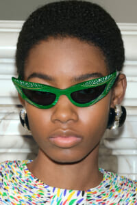 Young person wearing bright green rhinestone-embellished cat-eye sunglasses and colorful patterned top, bold fashion pose.