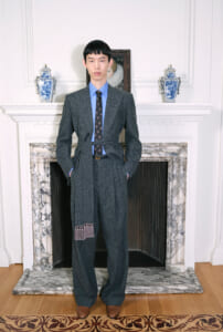 Young man in a charcoal textured suit with a blue shirt and patterned tie, standing with hands in his pockets in front of a marble fireplace.”,