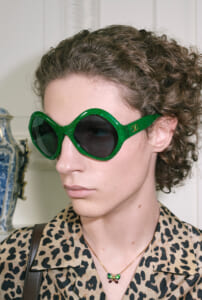 Person wearing oversized green sunglasses with dark lenses, a leopard-print shirt, and a butterfly-shaped green pendant necklace in a neutral indoor setting.