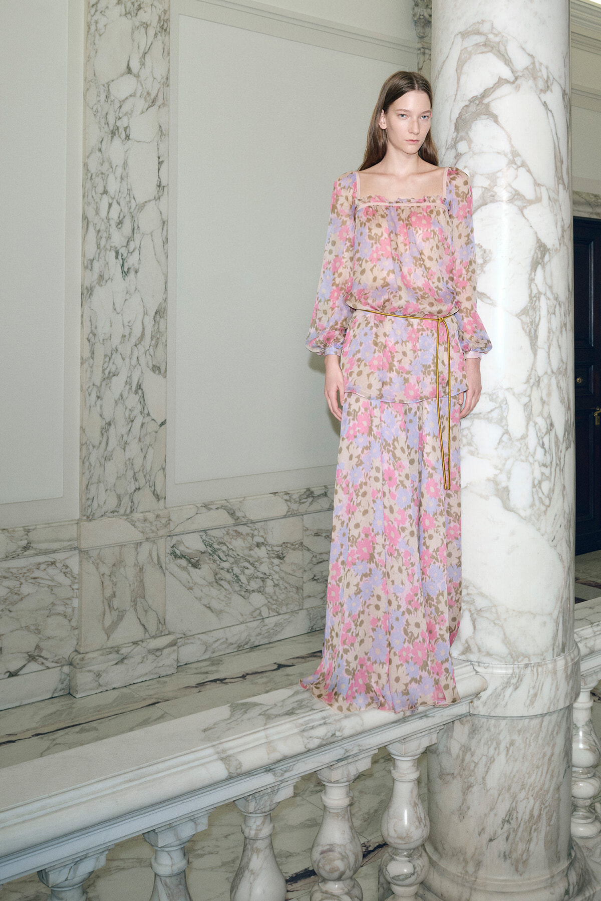 Woman in a pastel floral maxi dress posing on a marble balcony inside a marble-lined room.