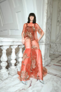 Model wearing a coral sheer embroidered gown, leaning on a marble railing in a grand marble hallway.