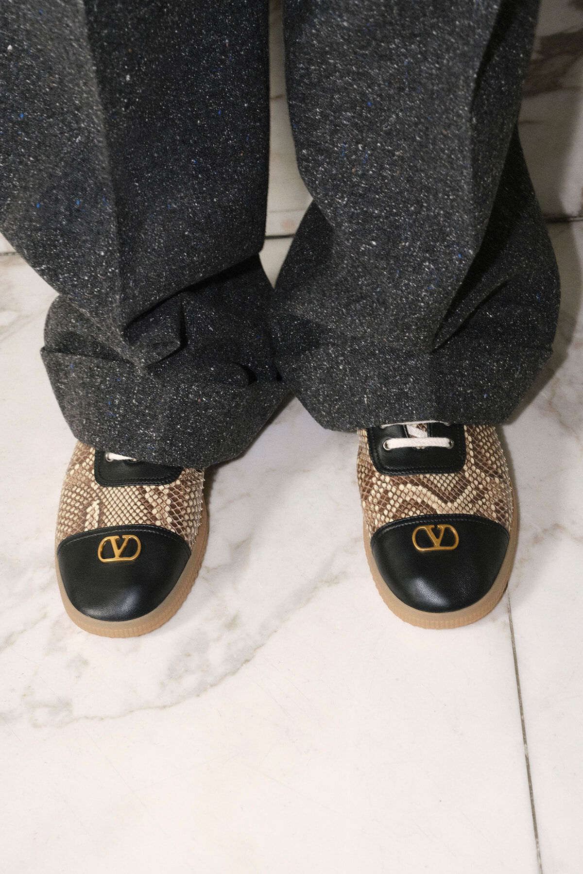 Person wearing glittery dark pants standing in snakeskin-pattern loafers with black toe caps and gold logo on a marble floor.