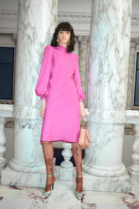Model wearing a bright pink midi dress with puffed sleeves, standing among marble columns, holding a blush handbag.