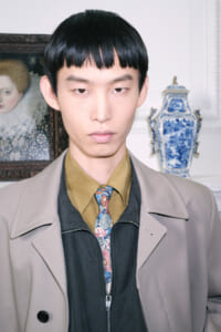 Portrait of a young man with straight black hair, wearing a beige coat over a black jacket and a floral tie, standing against a decorative background with a blue-and-white vase.