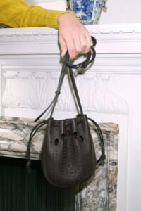Hand in a yellow sweater holding a brown leather drawstring handbag by its straps in front of a marble fireplace