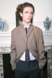 Male model in a taupe blazer, striped tie, white shirt, and dark trousers, with a tan shoulder bag, posed in a refined room with marble and blue ceramics.