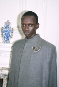 Portrait of a young Black man in a gray textured high-collar coat with a jeweled butterfly brooch, standing beside a blue-and-white porcelain vase.