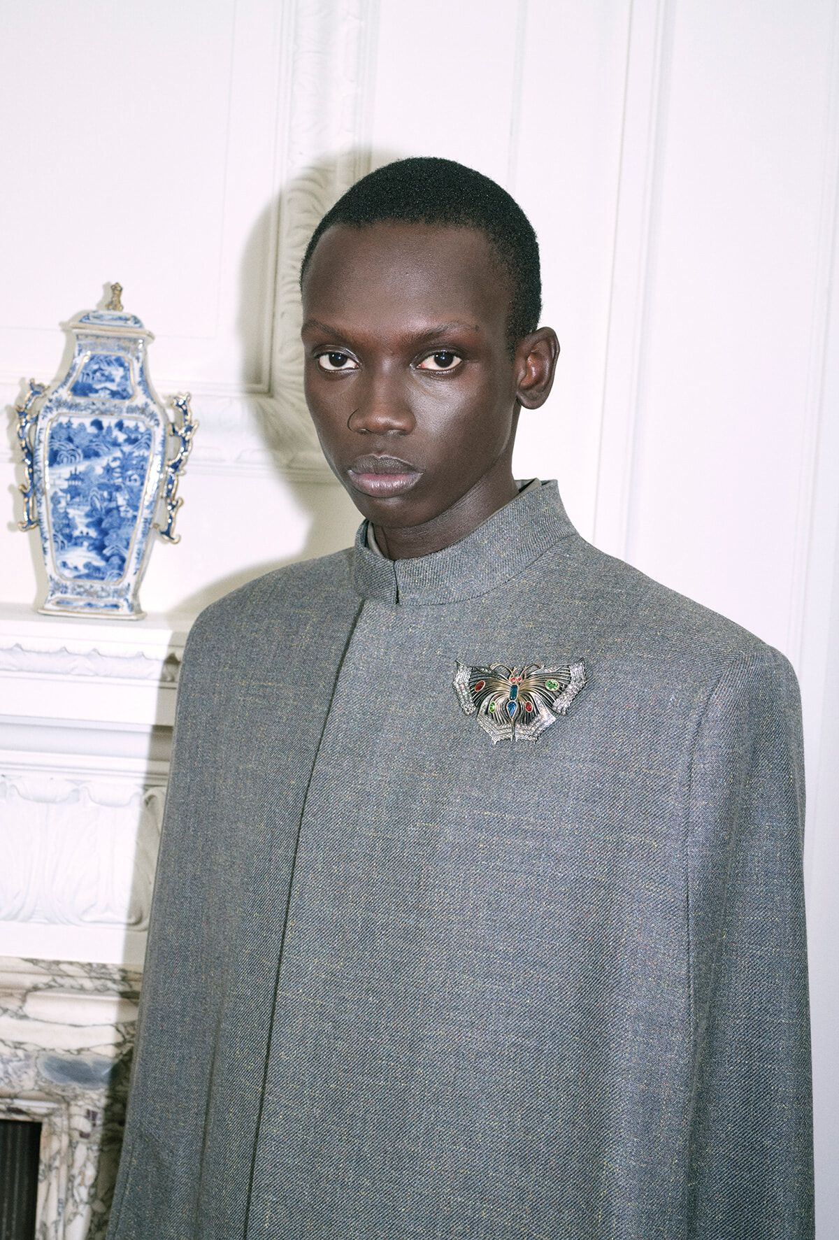Portrait of a young Black man in a gray textured high-collar coat with a jeweled butterfly brooch, standing beside a blue-and-white porcelain vase.