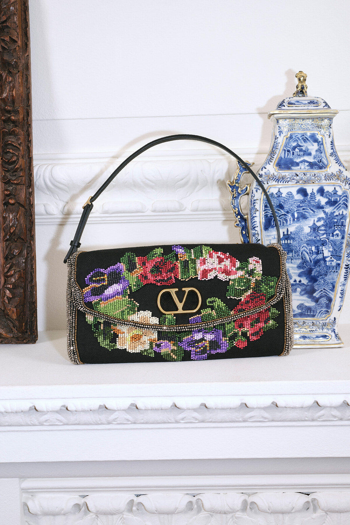 Beaded black handbag with colorful floral embroidery and a gold logo, resting on a white mantel beside a blue-and-white porcelain vase.