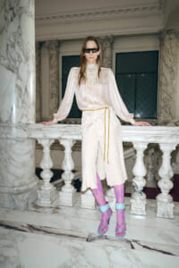 Fashion model in a cream, dotted sheer dress with a belt, standing at marble railing in a grand interior wearing purple tights and blue heels with sunglasses.