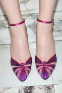Pair of pink and purple striped pointed-toe heels with bow knots and ankle straps on a marble floor.