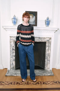 Person standing in a bright room in front of a marble fireplace, wearing a striped knit sweater and wide-leg blue jeans with sandals.