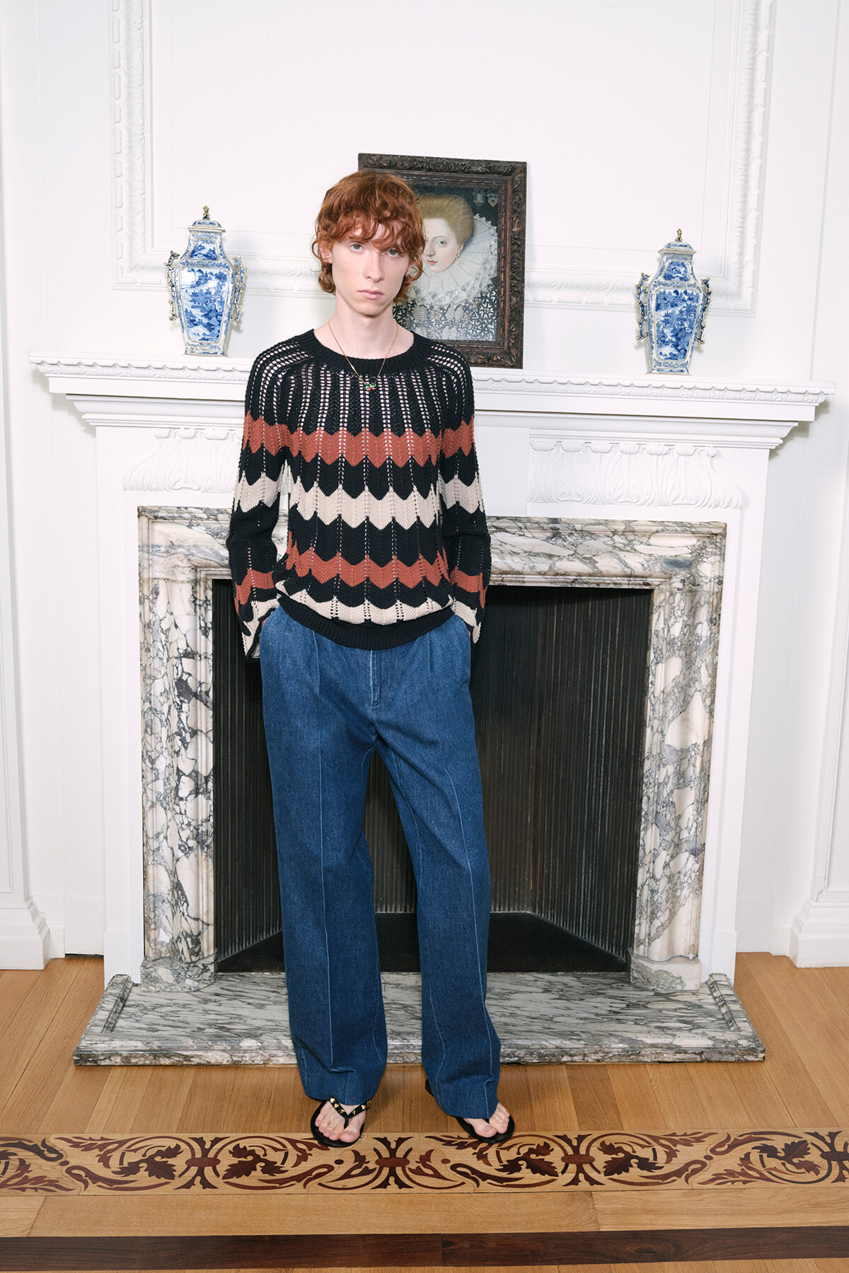 Person standing in a bright room in front of a marble fireplace, wearing a striped knit sweater and wide-leg blue jeans with sandals.