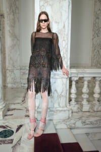 Model wearing a sheer black dress with multicolored bead fringe and sunglasses, standing in a marble hallway with pink tights and pink sandals.