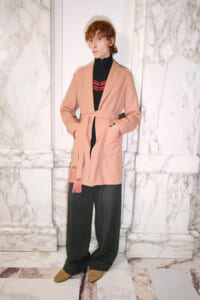 Person in a peach belted coat over a black zip top with red stripes, wearing wide charcoal trousers and tan woven shoes, standing against marble walls.