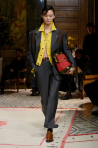Runway model in a gray suit with a bright yellow shirt, carrying a red handbag on the catwalk.