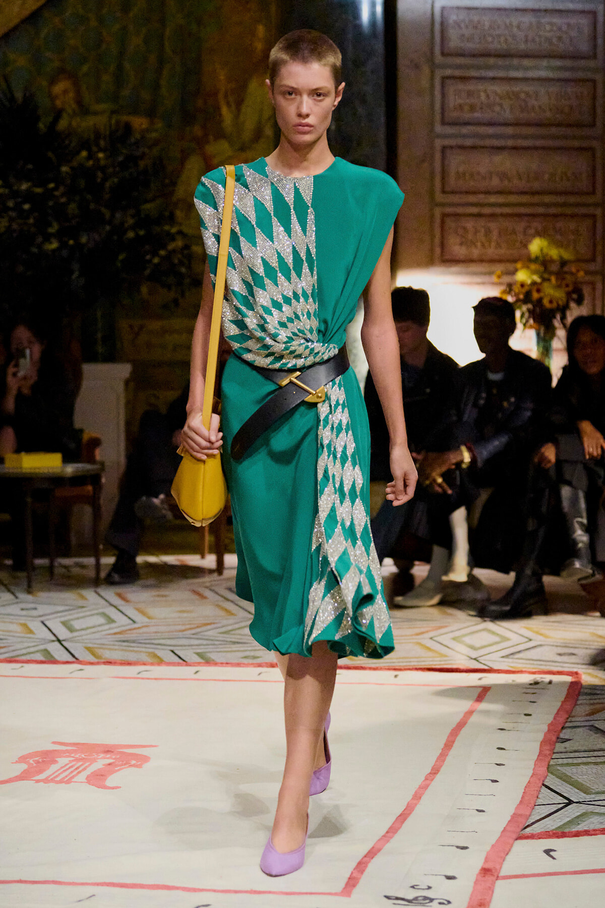 Model walks a teal-green dress with silver diamond pattern, belt, and a yellow crossbody bag on a fashion runway.