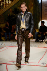 Model walking a fashion runway in a black quilted leather jacket, denim shirt, beige zip sweater, and dark brown leather pants; audience seated in the background.
