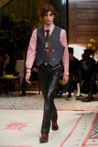 Male model walks a runway wearing a pink plaid shirt, dark vest with triangular pockets, black leather pants, and red shoes.