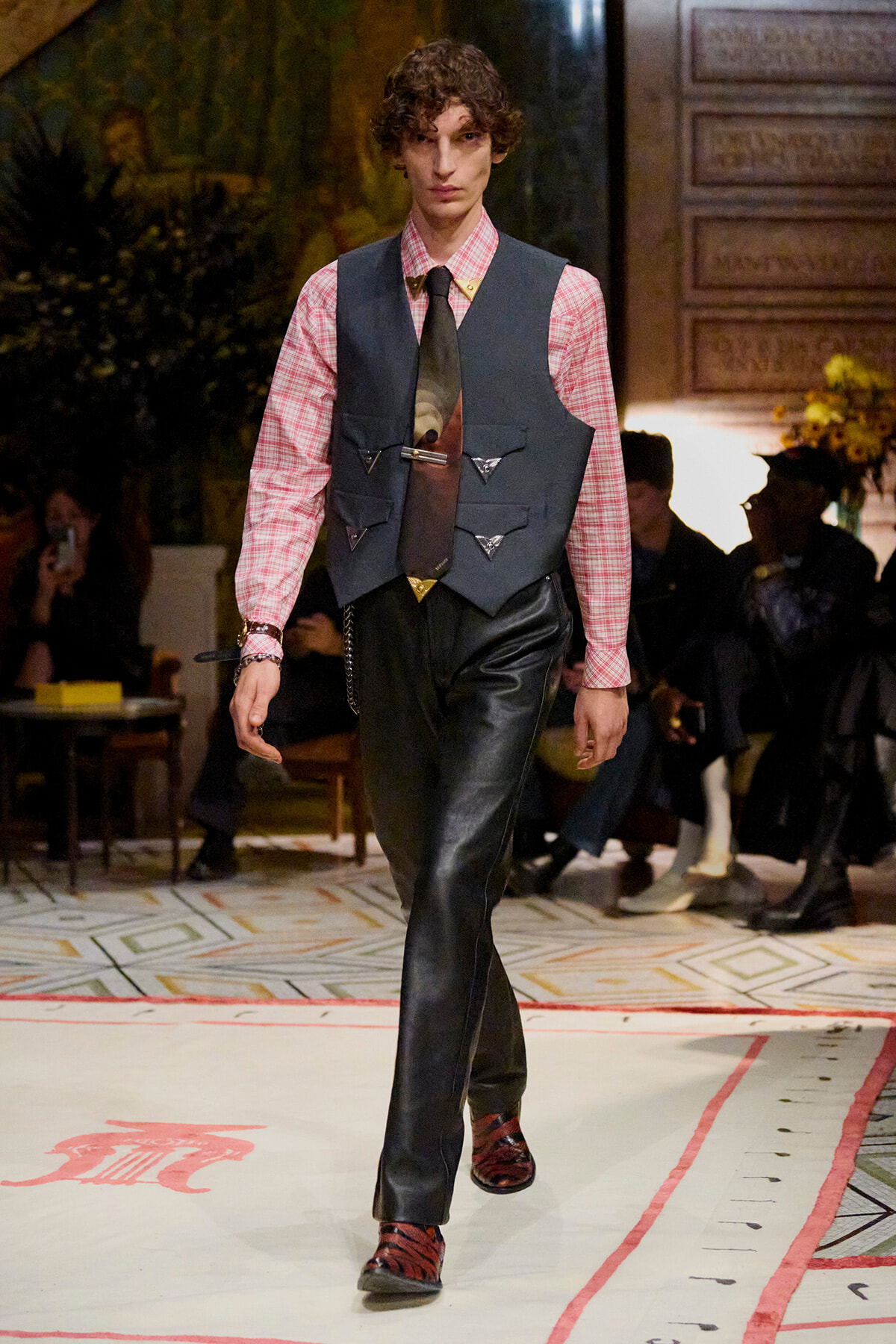 Male model walks a runway wearing a pink plaid shirt, dark vest with triangular pockets, black leather pants, and red shoes.