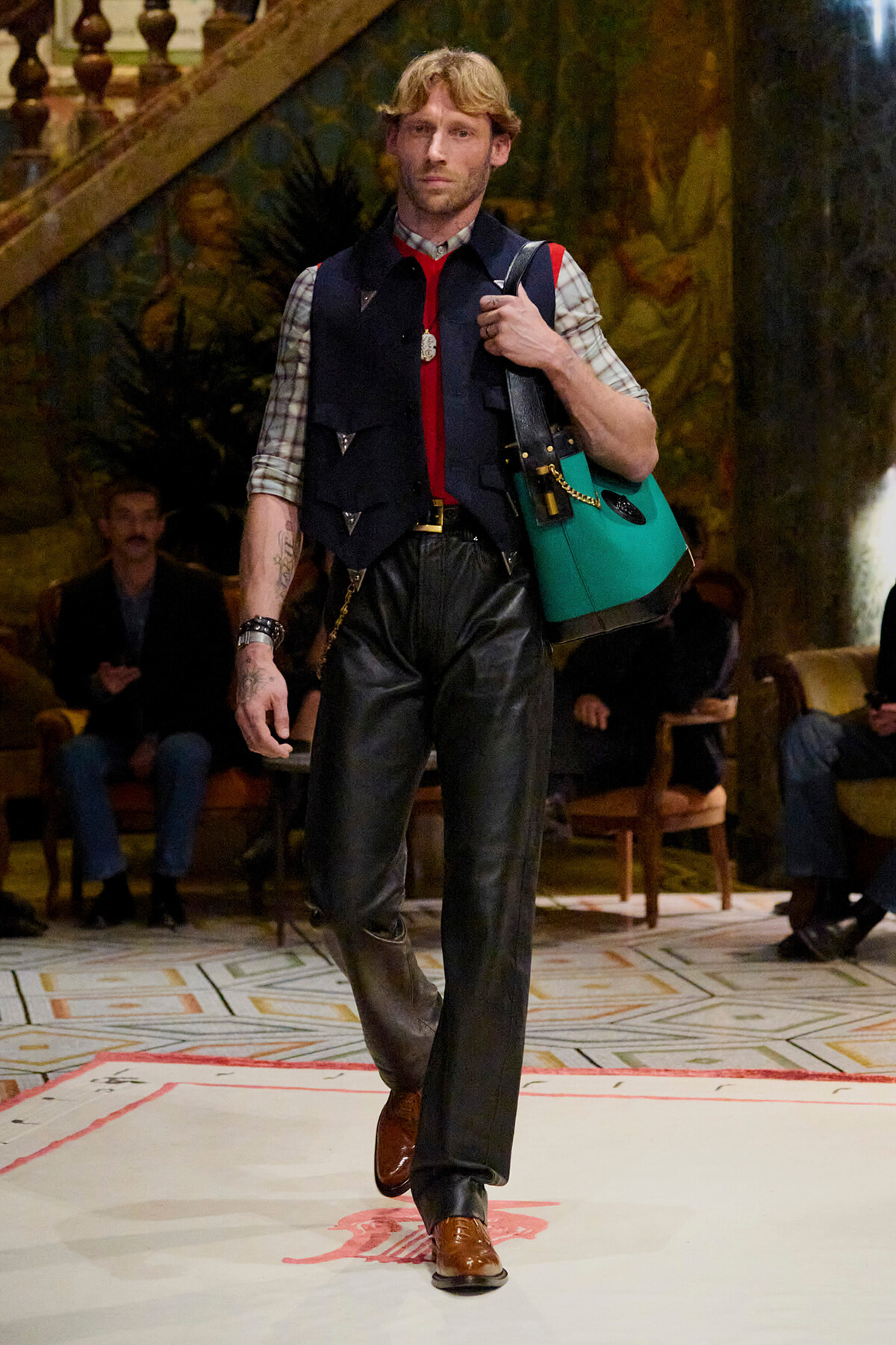 Male model walks a runway in a plaid shirt, navy vest, red tie, black leather pants, brown shoes, and a teal bag over the shoulder.