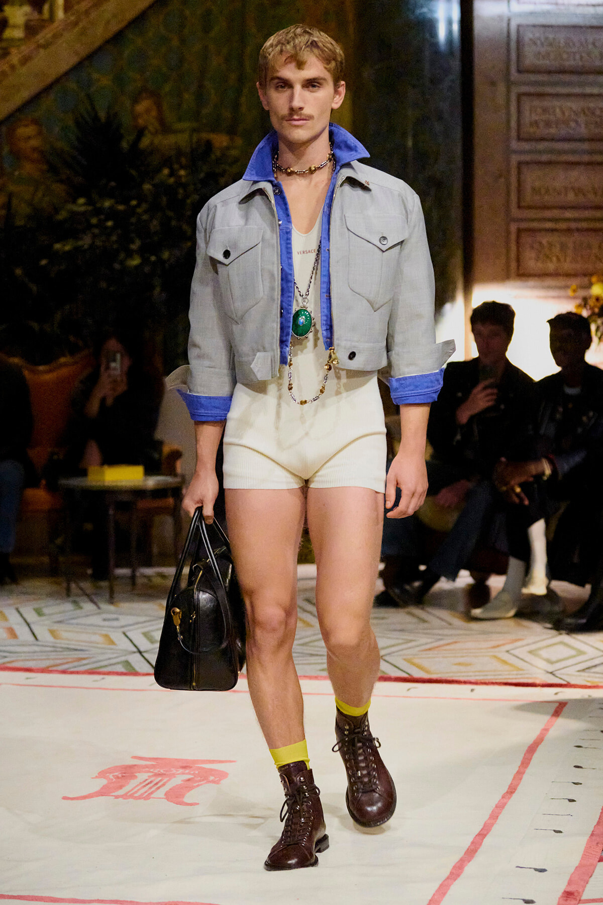 Male model on a runway wearing a light denim jacket over a white bodysuit, cream shorts, yellow socks, brown boots, and a black briefcase bag. Accessory necklace visible.