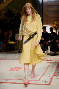 Model wearing a pale yellow midi dress with a metallic draped sash, wide belt, pink shoes, and a green shoulder bag walking a fashion runway.