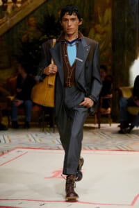 Male model walking a fashion runway in a dark blue suit with brown vest and turquoise shirt, carrying a tan shoulder bag and wearing brown boots.