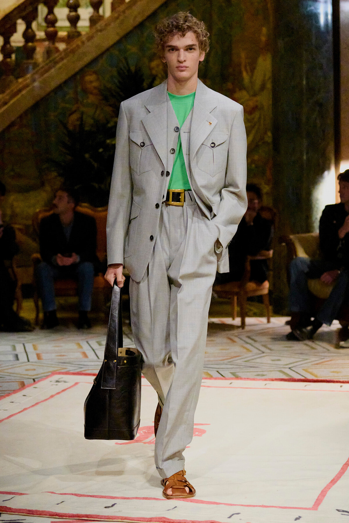 Male model on a runway wearing a pale gray suit with a bright green shirt, brown sandals, and a black leather tote bag.