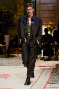 Male model walks a fashion runway in a color-blocked navy and black jacket over a green vest, pink shirt, black trousers, and chunky boots.