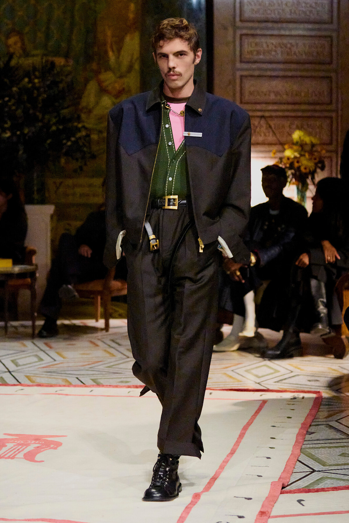 Male model walks a fashion runway in a color-blocked navy and black jacket over a green vest, pink shirt, black trousers, and chunky boots.
