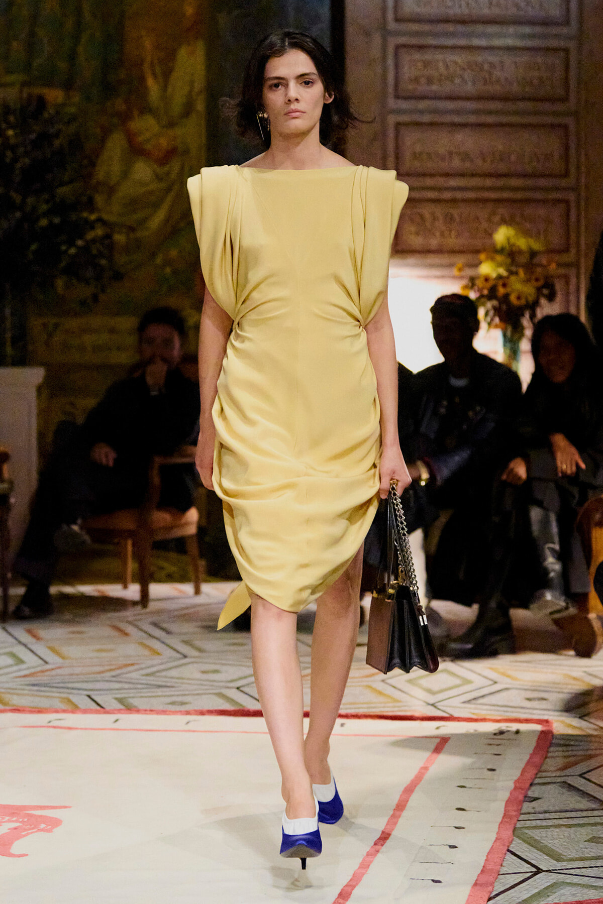 Model walking a runway in a pale yellow sculptural dress, carrying a small black handbag with a chain strap and blue-and-white heels.] ,