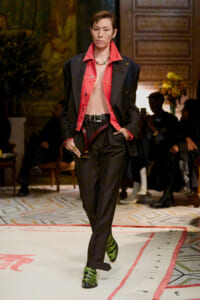 Model walking a runway in a red collared shirt under a black blazer, black trousers, and bright green zebra-print boots.