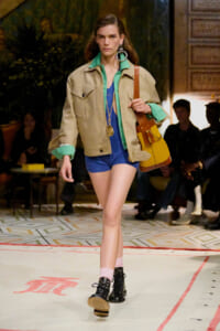 Model on a runway wearing a tan oversized jacket over a bright teal shirt and blue romper, with a yellow handbag and chunky boots.