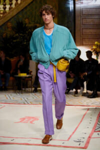 Male model walks a runway in a teal bomber jacket, blue shirt, lavender pants, brown loafers, yellow belt bag; audience in background.