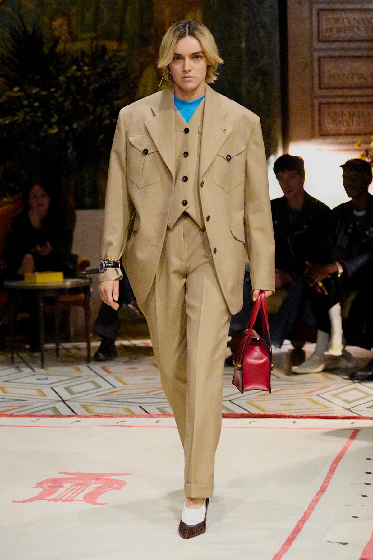 Model walking a runway in a tan suit with a blue top, carrying a red handbag; audience seated in the background.