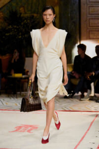 Model walks a runway in a cream draped V-neck dress, red and white heels, and a dark brown crocodile-embossed handbag with a chain strap.
