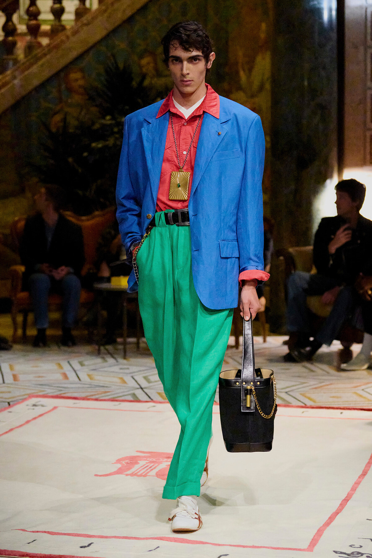 Male model walks the runway in a bright blue blazer, red shirt, green trousers, white sneakers, and carries a black handbag with a chain.