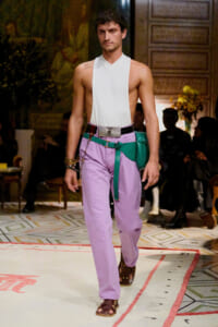 Male model walking a runway in a white sleeveless top and lavender pants with a green belt and turquoise pouch, during a fashion show.