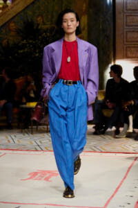 Model walks a fashion runway in a purple blazer, red top, and bright blue high-waisted pants, carrying a dark handbag.