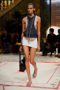 Model walking a runway in a navy sleeveless top with layered beige shirt, white mini skirt, and layered gold necklaces, carrying a black tote.