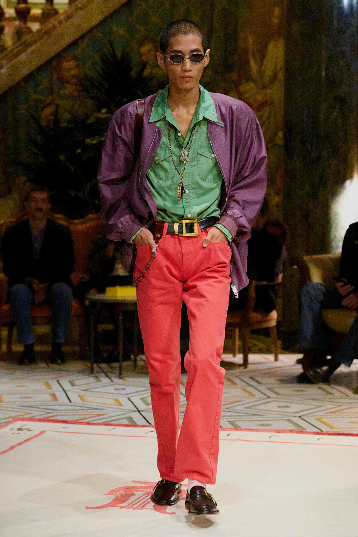 Model walking a runway in a green shirt, purple jacket, coral pink trousers, black loafers, and sunglasses, accessorized with gold chains and a belt.