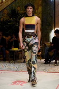 Male model walks a fashion runway wearing a sleeveless color-block top and jeans printed with vintage faces; audience seated in background.