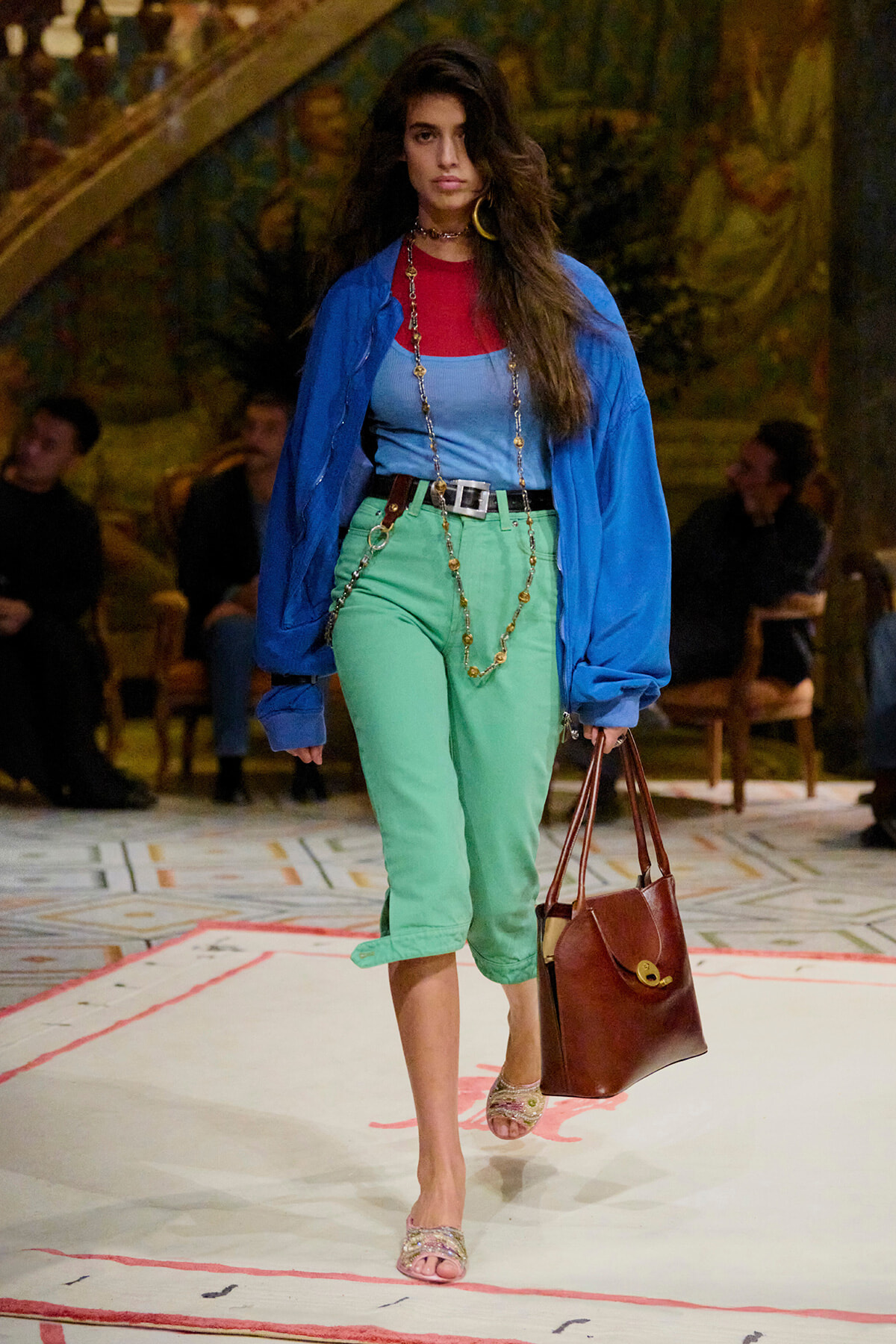 Model walking the runway in a blue oversized jacket, red top under a light blue tank, green capri pants, and a brown handbag.